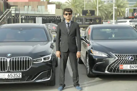A chauffeur in a dark suit between a black BMW and a black Lexus