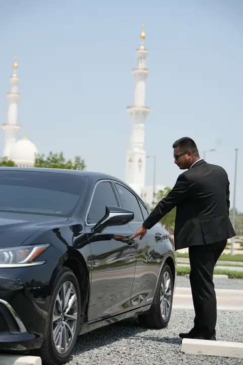 A chauffeur opening the rear door of a black Lexus, the Grand Mosque behind