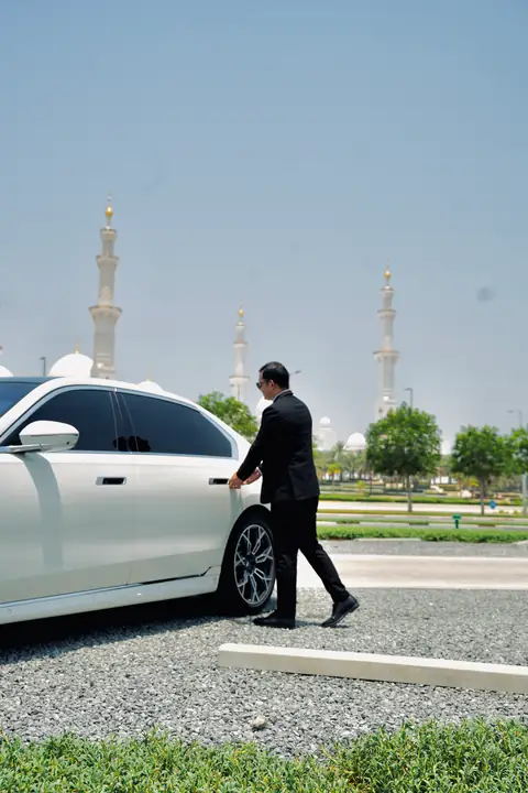 A chauffeur walking beside a white BMW 7 Series, the Grand Mosque behind