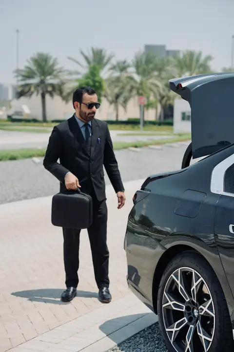 A chauffeur with a briefcase beside the open boot of a black car