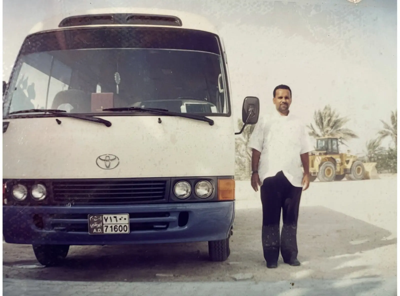 Aboobacker Thuppath in 1999, in a white shirt, standing beside the front of his first bus — a white Toyota Coaster on Abu Dhabi plates — with palm trees and a yellow loader behind him.