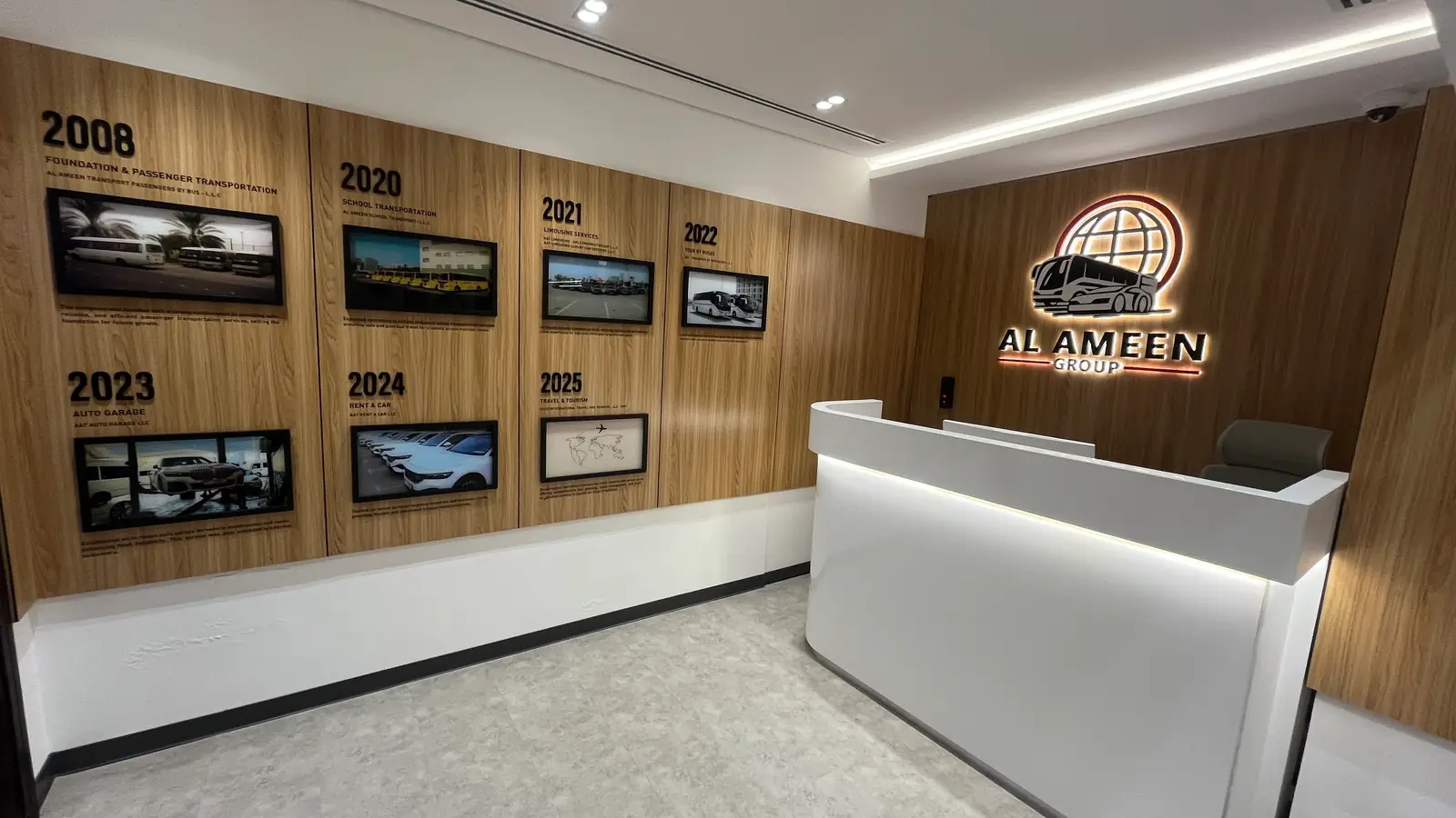 The reception of the group’s Dubai head office: a curved white desk, the timeline wall to the left, and the illuminated Al Ameen Group mark and name on wood panelling behind the desk.